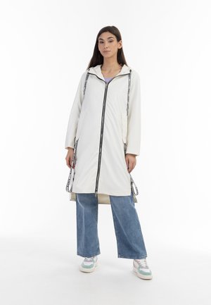 Giacca impermeabile bianca con chiusura a zip, cordini e due tasche frontali. Indossata sopra jeans a gamba larga blu e sneakers multicolori.
