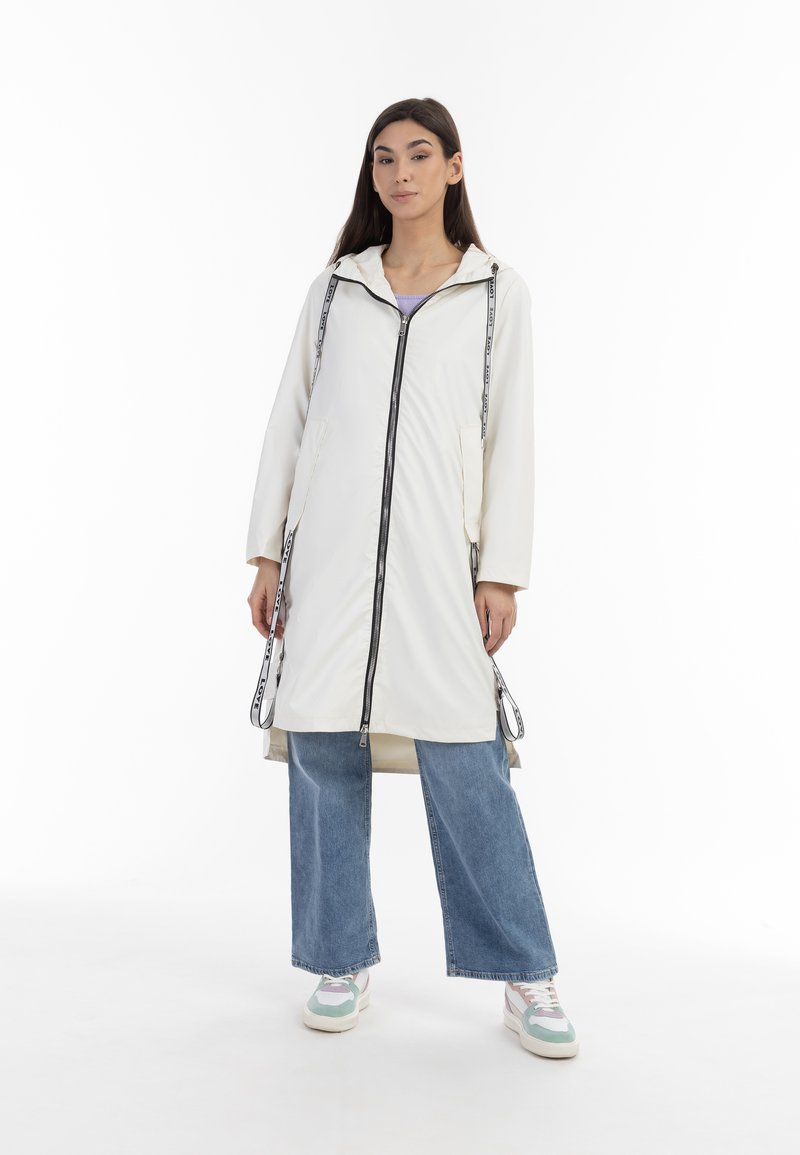 Chaqueta blanca impermeable con cierre de cremallera, cordones y dos bolsillos frontales. Lleva puesta sobre unos vaqueros anchos azules y zapatillas multicolores.