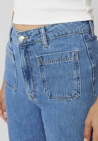 Jeans en denim bleu clair à taille haute, avec deux poches arrière, des coutures dorées et une fermeture à bouton en argent à la taille.