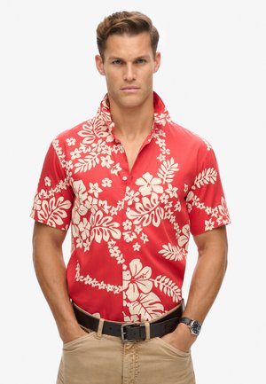Camisa roja de manga corta con patrones de flores y hojas blancas, diseño con cuello y cierre de botones. Se lleva con pantalones de color arena y un cinturón negro.