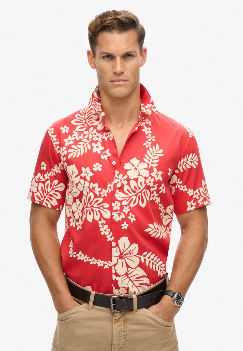Rood overhemd met korte mouwen met witte bloemen- en loofpatronen, met kraag en knoopsluiting. Draagt een zandkleurige broek en een zwarte riem.