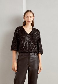 Schwarzes Paillettentop mit V-Ausschnitt, kurzen Ärmeln und oversized Schnitt, kombiniert mit schwarzen Lederhosen. Glatte Textur und auffälliger Glanz.