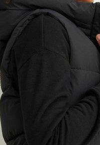 Czarna kamizelka puchowa z wysokim kołnierzem, z pikowanymi sekcjami i gładką fakturą materiału. Pod spodem znajduje się dopasowana czarna bluzka z długim rękawem.