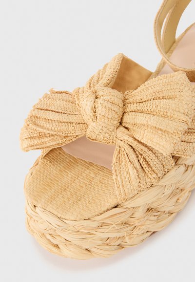 Woven straw platform sandal in beige with a large knotted bow detail on the front and a textured, braided jute sole. Smooth inner lining.