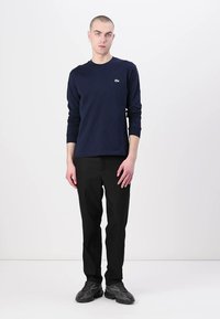 Chemise à manches longues navy avec col rond et logo sur la poitrine, associée à un pantalon droit noir et des baskets grises, sur fond blanc.