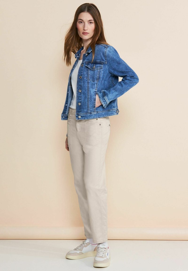 Jeansjacka i mellanblått med knäppning fram, bröstfickor och subtila blekningar. Beiga byxor och vita sneakers fulländar outfiten.