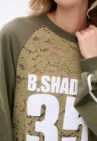 Olijfgroene sweatshirt met een kanten vooroverlay met bloemenpatroon, grote witte gedrukt cijfers en contrasterende ribgebreide halslijn en mouwen.