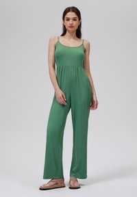 Combinaison verte côtelée avec des bretelles spaghetti et un buste ajusté, se déversant en pantalons larges. Portée avec des sandales plates marron.