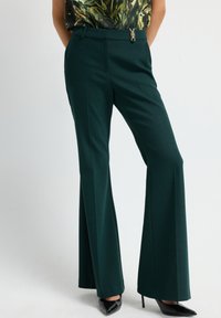 Vloeiende groene broek met een gladde textuur, voorzien van een decoratieve ritsaccent bij de taille en voorste plooien voor extra detail.