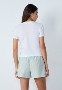 Camiseta blanca de manga corta exhibida con pantalones cortos de cuadros verdes. La tela parece suave, con un diseño simple y un ajuste relajado.