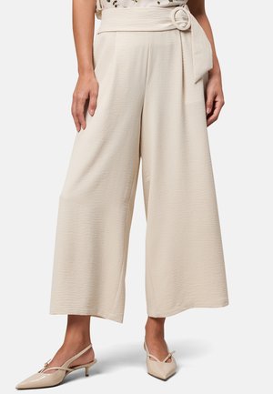 Beige wijde broek met een gestructureerde stof, voorzien van een hoge taille en een knoopdetail bij de riem. Gecombineerd met lichtgekleurde hakken.