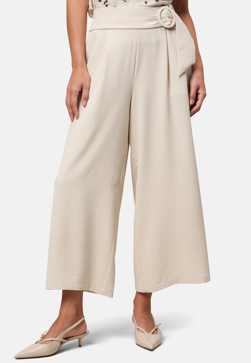 Beige wijde broek met een gestructureerde stof, voorzien van een hoge taille en een knoopdetail bij de riem. Gecombineerd met lichtgekleurde hakken.