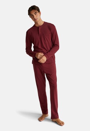 Joven de pie descalzo, vistiendo un conjunto de pantalones y una camisa de manga larga color burdeos a juego, con un bolsillo en el pecho, sobre un fondo blanco.