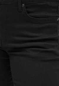 Gros plan sur un jean en denim noir montrant la poche avant, la boucle de ceinture, le bouton et la texture du tissu avec de légères plis.