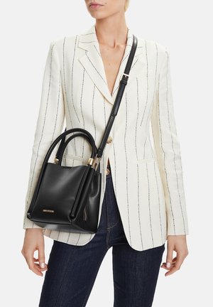 Frau in weißem Nadelstreifenblazer und dunklen Jeans, die eine schwarze Ledertasche mit Tragegriffen oben und Schulterriemen trägt.