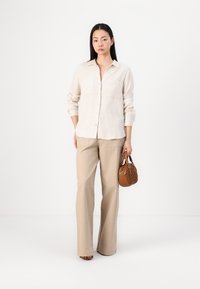 Lichtbeige linnen blouse met lange mouwen, twee voorzakken; gecombineerd met beige wijde broek en een kleine bruine handtas.