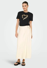 Camiseta negra con un diseño de corazón dorado, combinada con una falda envolvente de crema hasta la rodilla y chanclas de plataforma negras. Textura de tejido suave.