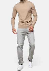 Beige coltrui met geribde boorden, gecombineerd met lichtgrijze spijkerbroek en witte sneakers. Casual, slim fit ontwerp met minimale details.