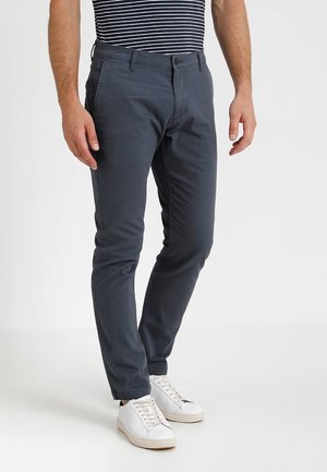 Mann trägt dunkelgraue, schmal geschnittene Hose und weiße Sneakers, steht vor weißem Hintergrund, gestreiftes Hemd teilweise sichtbar.