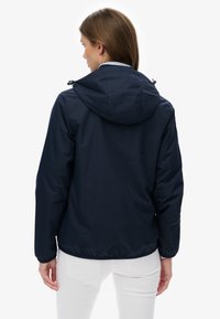 Femme aux cheveux bruns portant une veste à capuche bleu marine et un pantalon blanc, tournée de dos sur un fond blanc uni.