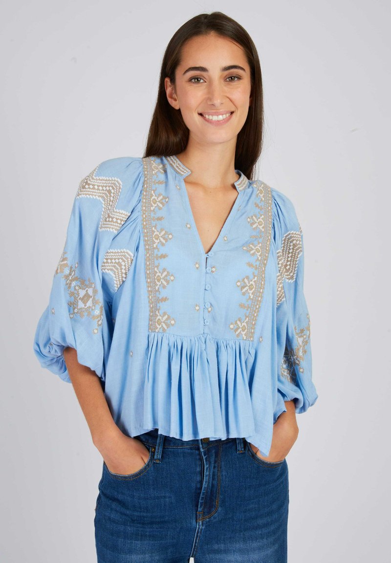 Lichtblauwe blouse met pofmouwen, geborduurde gouden patronen, V-hals en knopen aan de voorkant; geplooide zoom en comfortabele pasvorm.