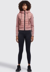 Gepolsterte rosa Jacke mit Kapuze, schwarzen Reißverschlüssen und gerippten Bündchen, kombiniert mit schwarzen Leggings und schwarzen Sportschuhen.