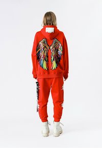 Sudadera con capucha roja con diseño de alas de lentejuelas coloridas en la parte trasera, combinada con pantalones joggers a juego que presentan detalles desgastados y detalles audaces en blanco.