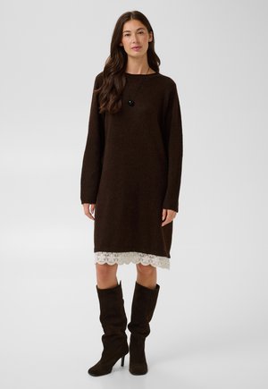 Femme aux longs cheveux foncés portant une robe-pull marron à manches longues avec un ourlet en dentelle blanche et des bottines marron foncé à talons larges.