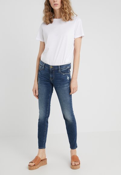 Jean skinny bleu avec des détails déchirés, associé à un t-shirt blanc et des sandales à talons compensés en liège tan. Tissu lisse avec une silhouette ajustée.