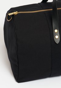 Černá plátěná sportovní taška se zlatým zipem, koženými rukojeťmi a zlatými kovovými detaily. Měkká textilie a zaoblený tvar.