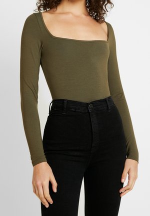 Vrouw draagt een olijfgroene top met lange mouwen en een vierkante halslijn, gecombineerd met een zwarte, hooggesneden spijkerbroek, tegen een effen achtergrond.