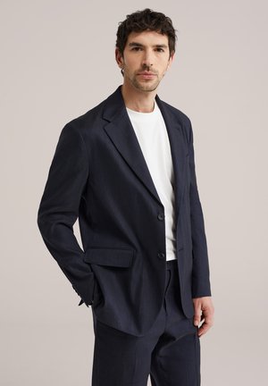 Homme aux cheveux bouclés foncés portant une veste de costume bleu marine, un pantalon assorti et un t-shirt blanc, debout devant un fond clair uni.