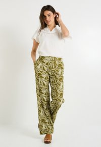 Blouse blanche à manches courtes avec un col, associée à un pantalon ample vert olive doté d'un imprimé botanique blanc et de poches latérales. Sandales à talons brunes.