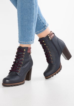 Bottes chevilles à talons hauts bleu marine avec lacets bordeaux et fermeture éclair, portées avec un jean bleu clair raccourci sur fond blanc.