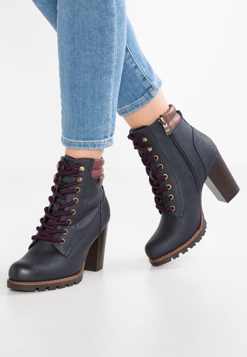 Bottes chevilles à talons hauts bleu marine avec lacets bordeaux et fermeture éclair, portées avec un jean bleu clair raccourci sur fond blanc.