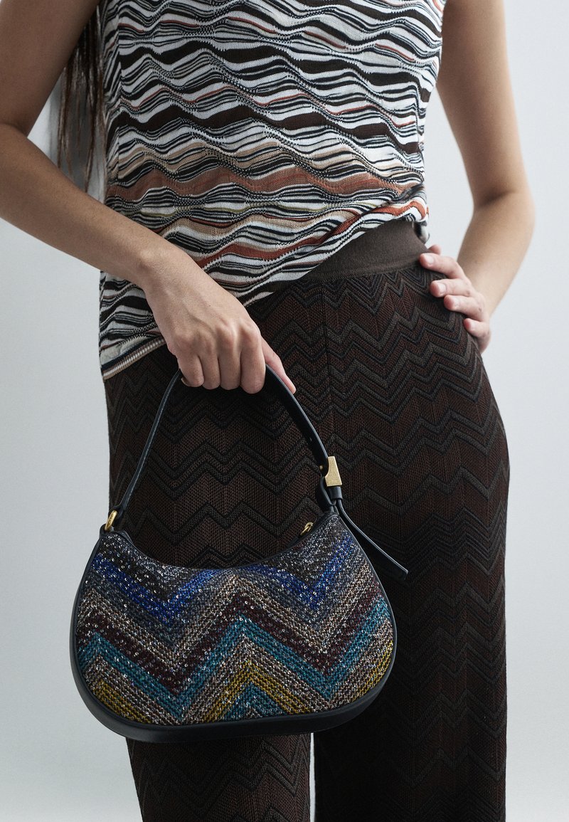 Kvinna som håller en liten handväska med flerfärgad zigzag-mönster, iklädd bruna byxor med zigzag-mönster och en randig ärmlös topp med vågor.