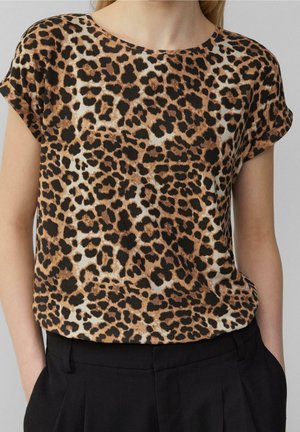 Kobieta w bluzce z krótkim rękawem w panterkę, wpuszczonej w czarne spodnie z wysokim stanem, ręce w kieszeniach, neutralne tło.