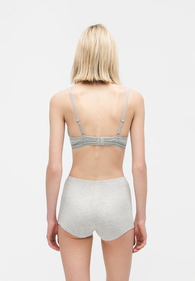 Kvinde med blondt hår iført en grå bh og matchende højtaljede shorts, stående med ryggen mod en ensfarvet baggrund.