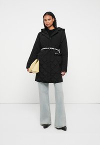 Cappotto trapuntato nero con cappuccio, dotato di una cintura bianca con logo, tasche frontali e una vestibilità rilassata. Abbinato a jeans a zampa d'elefante blu chiaro e una borsa gialla.