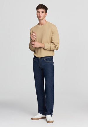 Maglietta a maniche lunghe beige a coste, jeans dritti blu scuro, sneakers bianche con dettagli marroni; modello in piedi davanti a uno sfondo chiaro.