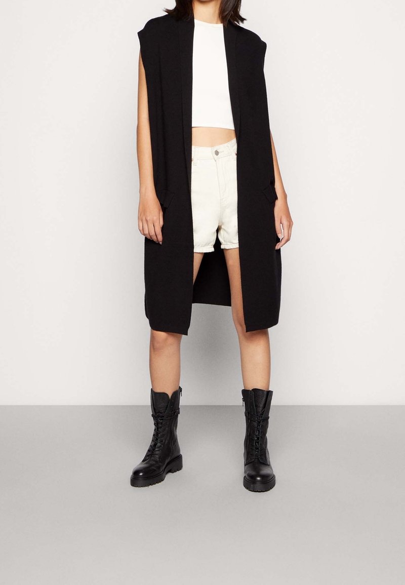 Femme portant un long gilet noir sans manches par-dessus un crop top blanc et un short blanc, assorti à des bottes de combat noires à lacets, debout.