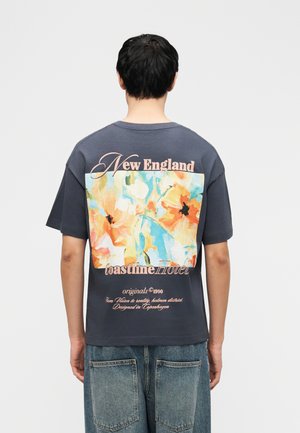 Personne aux cheveux foncés portant un T-shirt gris foncé avec un large motif floral coloré et le texte « New England coastline Hotel » au dos.