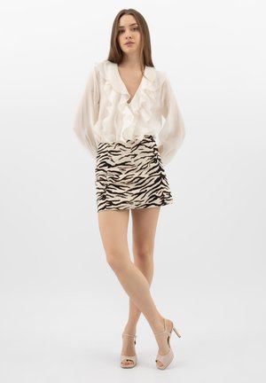 Jonge vrouw die een wit blouse met ruches draagt, een minirok met zebraprint en beige sandalen met hoge hakken, staand met gekruiste benen tegen een witte achtergrond.