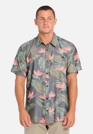 Uomo che indossa una camicia a maniche corte con bottoni, con motivo di foglie e fiori tropicali verdi e rosa su sfondo grigio.