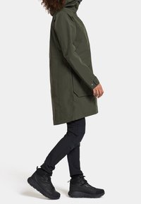 Olivgrüne wasserfeste Jacke mit Kapuze, gerade geschnitten, mit Seitentaschen und Druckknöpfen. Tragen mit schwarzen Hosen und Stiefeln.