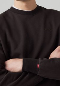 Felpa nera in cotone, con scollo a girocollo a costine e un discreto logo Levi's. Polsini con dettaglio etichetta rossa. Texture morbida.