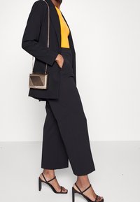 Blazer noir ajusté, top jaune cintré, pantalon noir taille haute, mini sac à fermoir doré, et sandales à talons noires avec lanières.