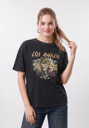 Νεαρή γυναίκα με μακριά μαλλιά, φορώντας κρίκους στα αυτιά, μαύρο μπλουζάκι με στάμπα "Los Angeles Wild Soul" με κεφάλια λεοπάρδαλης και μπλε τζιν.
