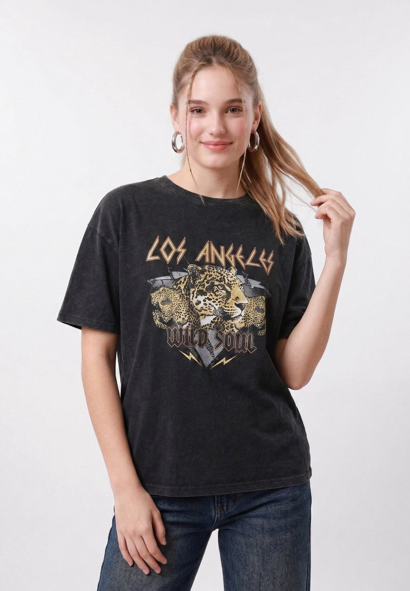 Νεαρή γυναίκα με μακριά μαλλιά, φορώντας κρίκους στα αυτιά, μαύρο μπλουζάκι με στάμπα "Los Angeles Wild Soul" με κεφάλια λεοπάρδαλης και μπλε τζιν.