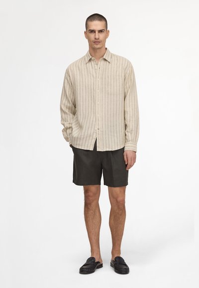 Homme debout portant une chemise beige à rayures verticales à manches longues, un short noir et des chaussures noires sans lacets sur fond blanc.